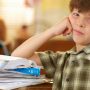 A young boy resting his chin on his hand while sitting at a desk covered with papers.