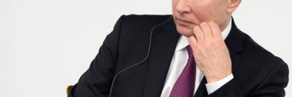 A man in a black suit and pink tie seated with a thoughtful expression.