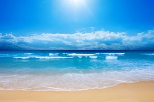 Gentle ocean waves lapping against a sandy beach under a clear blue sky with clouds.