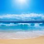 Gentle ocean waves lapping against a sandy beach under a clear blue sky with clouds.