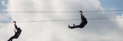 Two individuals ziplining across a cable with clouds in the background.