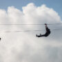 Two individuals ziplining across a cable with clouds in the background.