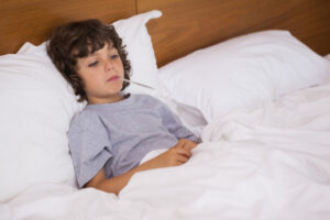 A young boy lying in bed with a thermometer in his mouth, looking unwell.
