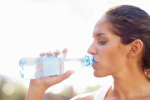Woman taking a sip from a clear plastic water bottle outdoors in natural light.