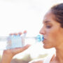 Woman taking a sip from a clear plastic water bottle outdoors in natural light.